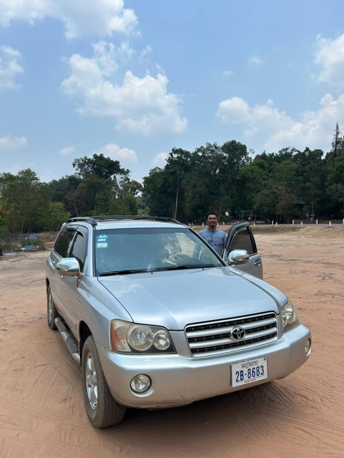 Smiling man stands by a silver Toyota SUV parked on a dirt lot near trees.