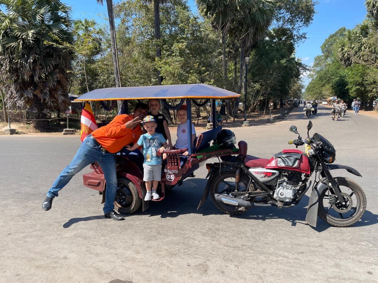 Man in orange shirt and children pose with a red tuk-tuk on a palm-lined road.